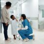 A young Black boy in a hospital gown stands beside his mother while interacting with a healthcare professional in scrubs.