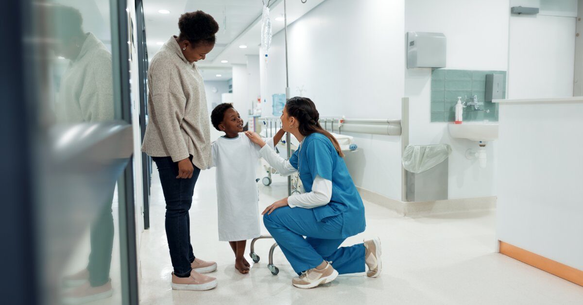 A young Black boy in a hospital gown stands beside his mother while interacting with a healthcare professional in scrubs.