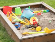 A children's sandbox filled with shovels, tools, toys, and games surrounded by a green grass backyard.