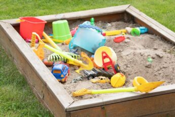 A children's sandbox filled with shovels, tools, toys, and games surrounded by a green grass backyard.