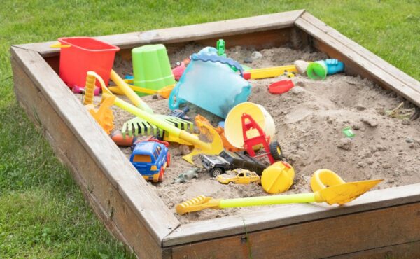 A children's sandbox filled with shovels, tools, toys, and games surrounded by a green grass backyard.