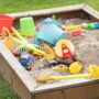 A children's sandbox filled with shovels, tools, toys, and games surrounded by a green grass backyard.