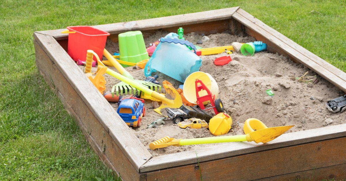 A children's sandbox filled with shovels, tools, toys, and games surrounded by a green grass backyard.