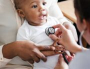 A person leaning forward examines a smiling black child sitting on an adult's lap with a stethoscope on the chest.