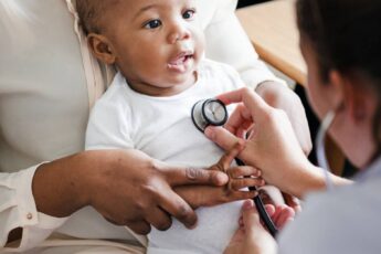 A person leaning forward examines a smiling black child sitting on an adult's lap with a stethoscope on the chest.