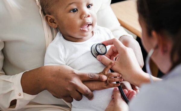 A person leaning forward examines a smiling black child sitting on an adult's lap with a stethoscope on the chest.