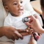 A person leaning forward examines a smiling black child sitting on an adult's lap with a stethoscope on the chest.