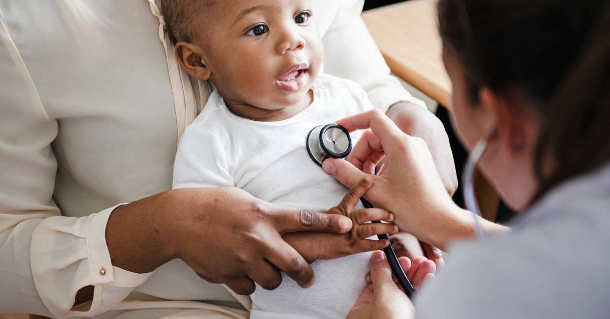 A person leaning forward examines a smiling black child sitting on an adult's lap with a stethoscope on the chest.