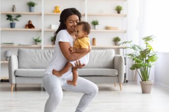 A Black mother smiles while holding her baby and doing a squat on a yoga mat in her bright, open living room.