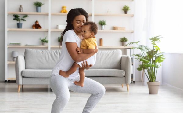 A Black mother smiles while holding her baby and doing a squat on a yoga mat in her bright, open living room.