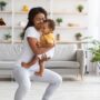 A Black mother smiles while holding her baby and doing a squat on a yoga mat in her bright, open living room.