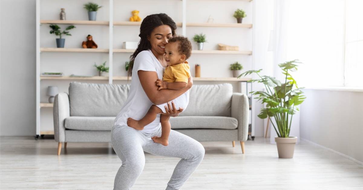 A Black mother smiles while holding her baby and doing a squat on a yoga mat in her bright, open living room.