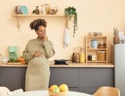 A black pregnant woman in a dress stands in her kitchen leaning against a counter while eating a snack.