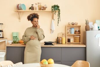 A black pregnant woman in a dress stands in her kitchen leaning against a counter while eating a snack.