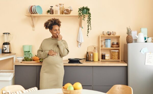 A black pregnant woman in a dress stands in her kitchen leaning against a counter while eating a snack.