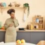 A black pregnant woman in a dress stands in her kitchen leaning against a counter while eating a snack.