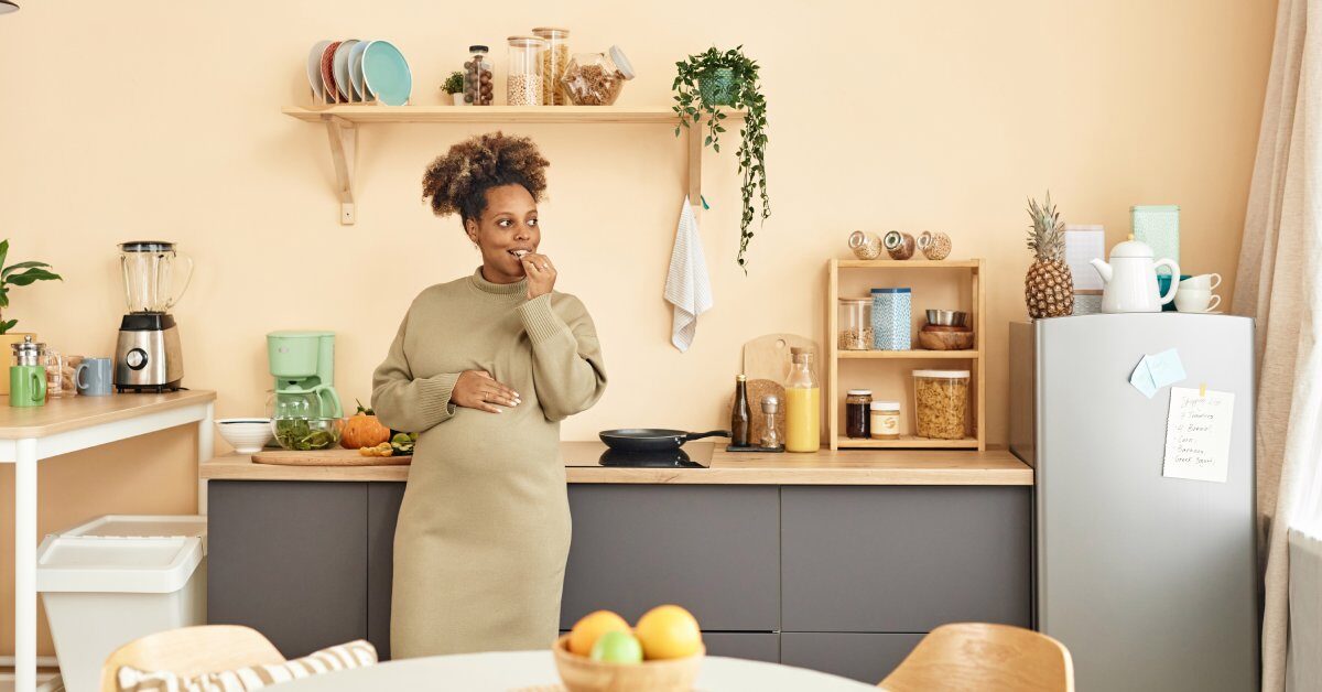 A black pregnant woman in a dress stands in her kitchen leaning against a counter while eating a snack.