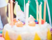 A young boy in a sleeveless green hoodie and yellow party hat opens his mouth to blow out candles on a cake.