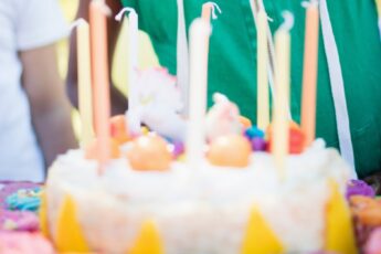 A young boy in a sleeveless green hoodie and yellow party hat opens his mouth to blow out candles on a cake.