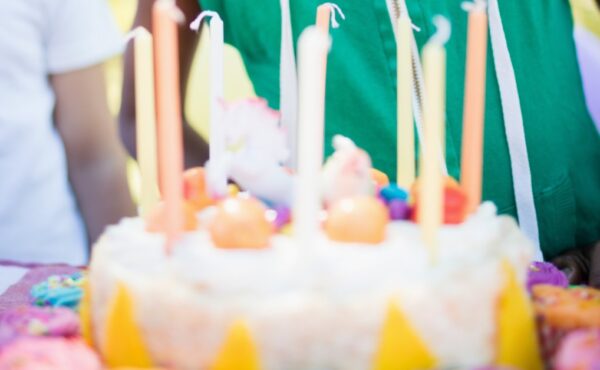 A young boy in a sleeveless green hoodie and yellow party hat opens his mouth to blow out candles on a cake.