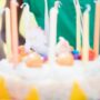 A young boy in a sleeveless green hoodie and yellow party hat opens his mouth to blow out candles on a cake.