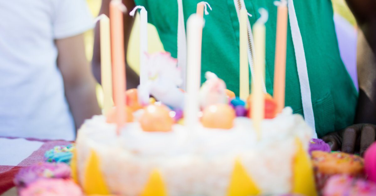 A young boy in a sleeveless green hoodie and yellow party hat opens his mouth to blow out candles on a cake.