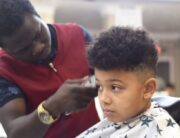 A boy with textured hair getting a haircut in a salon. The hairstylist is using an electric razor to shave the boy's head.