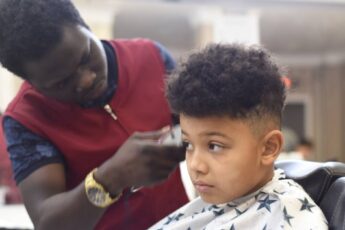 A boy with textured hair getting a haircut in a salon. The hairstylist is using an electric razor to shave the boy's head.