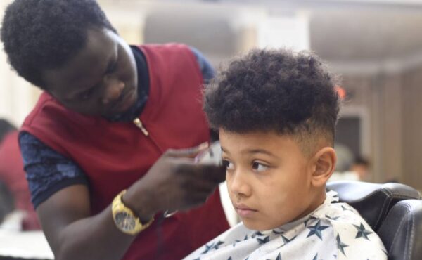 A boy with textured hair getting a haircut in a salon. The hairstylist is using an electric razor to shave the boy's head.