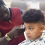 A boy with textured hair getting a haircut in a salon. The hairstylist is using an electric razor to shave the boy's head.