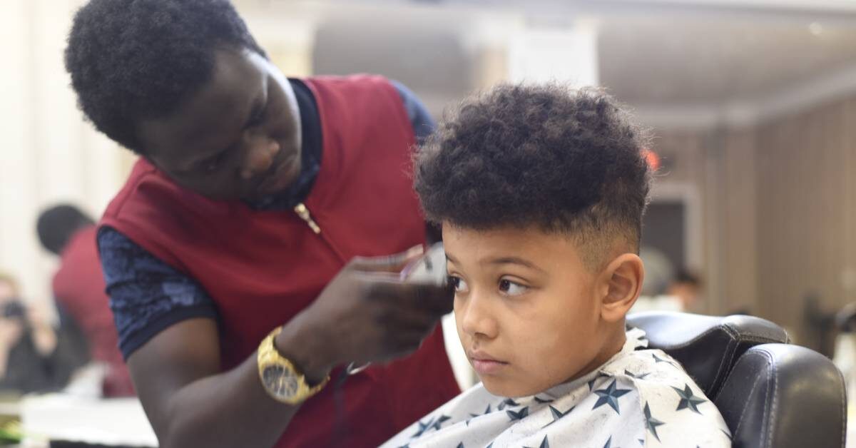 A boy with textured hair getting a haircut in a salon. The hairstylist is using an electric razor to shave the boy's head.