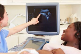 A pregnant woman lying on her back while another woman performs an ultrasound. Both are looking at the machine's screen.