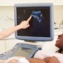 A pregnant woman lying on her back while another woman performs an ultrasound. Both are looking at the machine's screen.