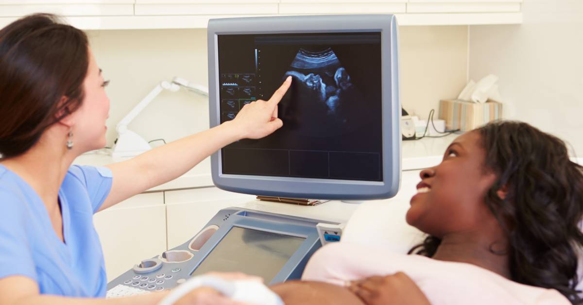 A pregnant woman lying on her back while another woman performs an ultrasound. Both are looking at the machine's screen.