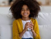 A young black girl with natural hair in a bright yellow cardigan is sitting on a white couch, smiling with a glass of water.