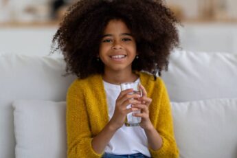 A young black girl with natural hair in a bright yellow cardigan is sitting on a white couch, smiling with a glass of water.