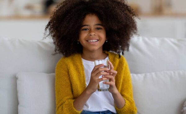 A young black girl with natural hair in a bright yellow cardigan is sitting on a white couch, smiling with a glass of water.