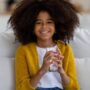 A young black girl with natural hair in a bright yellow cardigan is sitting on a white couch, smiling with a glass of water.