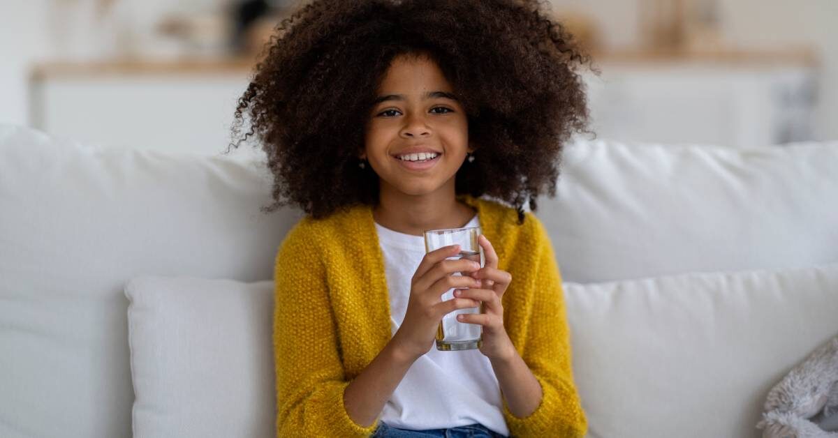 A young black girl with natural hair in a bright yellow cardigan is sitting on a white couch, smiling with a glass of water.