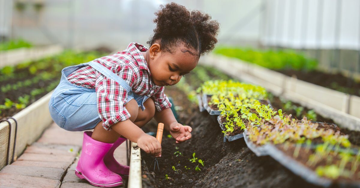 A young child in overalls and rain boots crouching by a planter bed and using a hand rake to dig in the dirt.