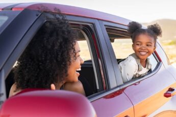 A Black mother and her young daughter in their red car. They're looking at each other from their windows.