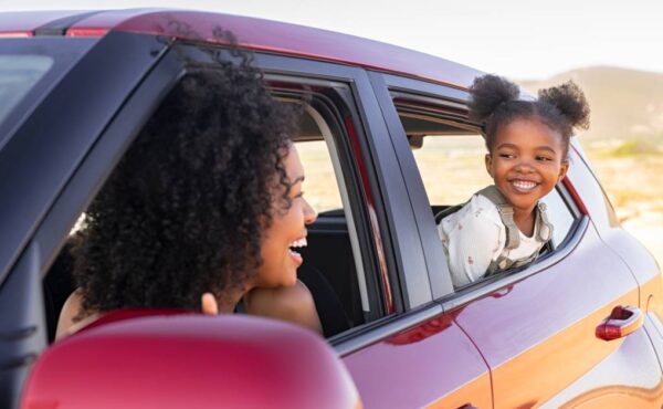 A Black mother and her young daughter in their red car. They're looking at each other from their windows.