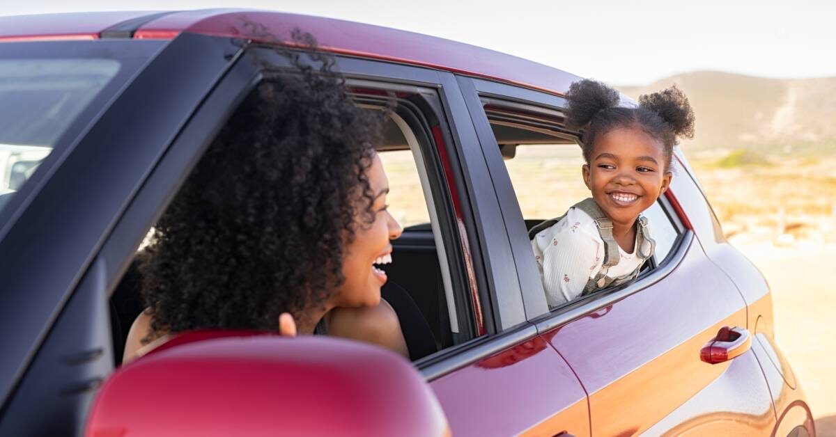 A Black mother and her young daughter in their red car. They're looking at each other from their windows.