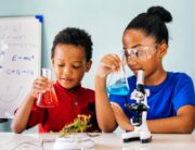 Two young children stand at a large white table holding glass beakers filled with different colored liquid.