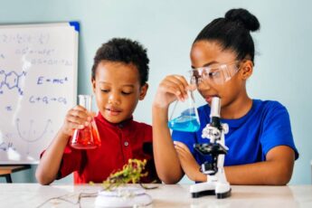 Two young children stand at a large white table holding glass beakers filled with different colored liquid.