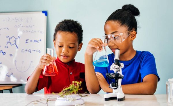 Two young children stand at a large white table holding glass beakers filled with different colored liquid.