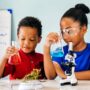 Two young children stand at a large white table holding glass beakers filled with different colored liquid.