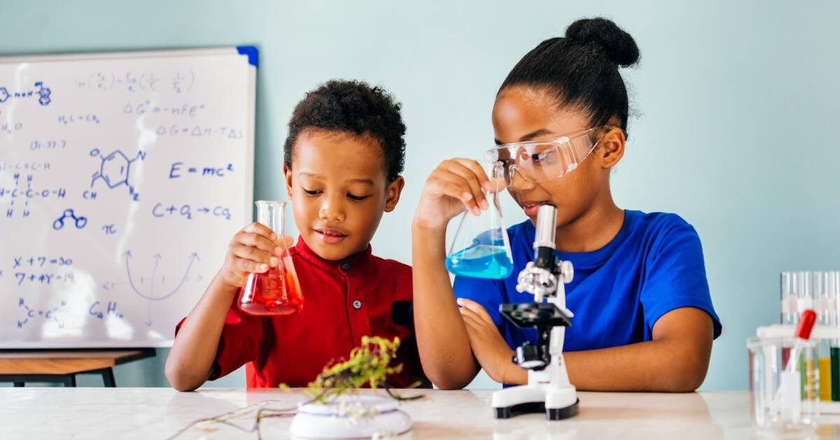 Two young children stand at a large white table holding glass beakers filled with different colored liquid.