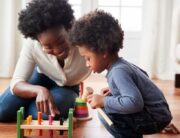 A mom and son playing with a wooden block toy on the floor of their home. The little boy is crouched and looking at the toy.