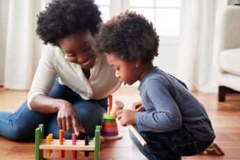 A mom and son playing with a wooden block toy on the floor of their home. The little boy is crouched and looking at the toy.